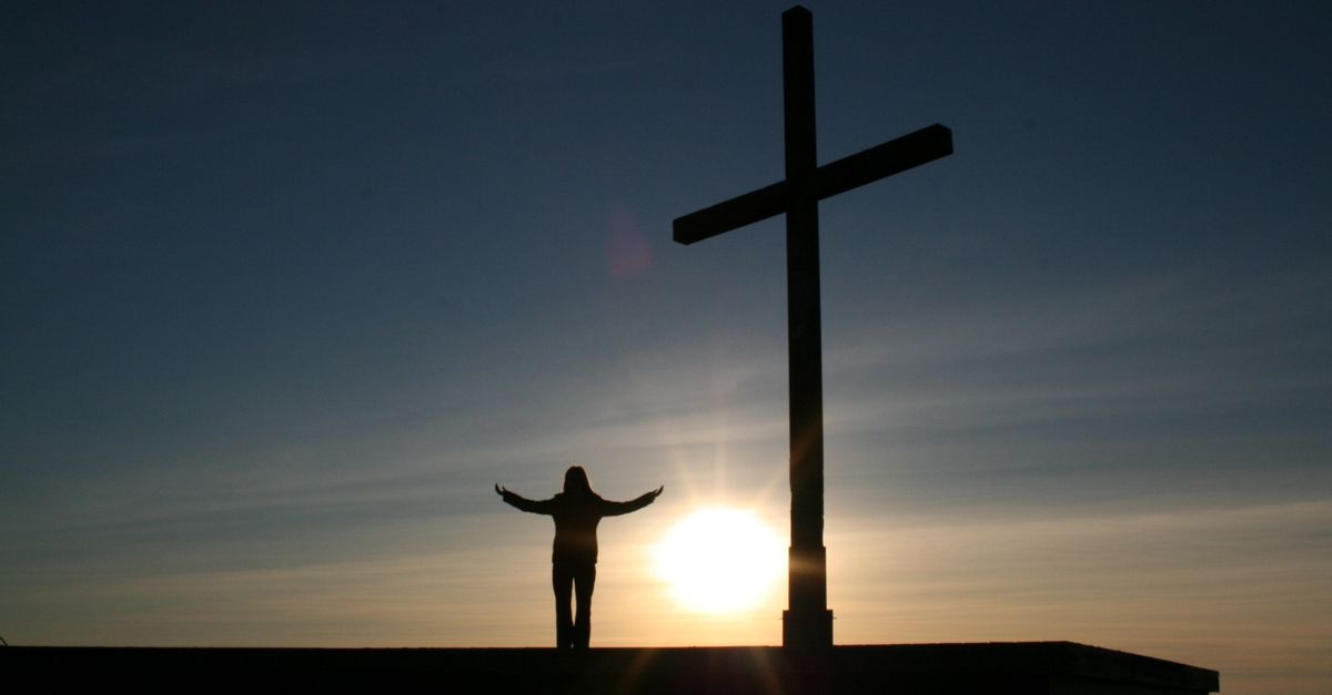 A person with open arms beside a cross at sunset, symbolizing faith and trust